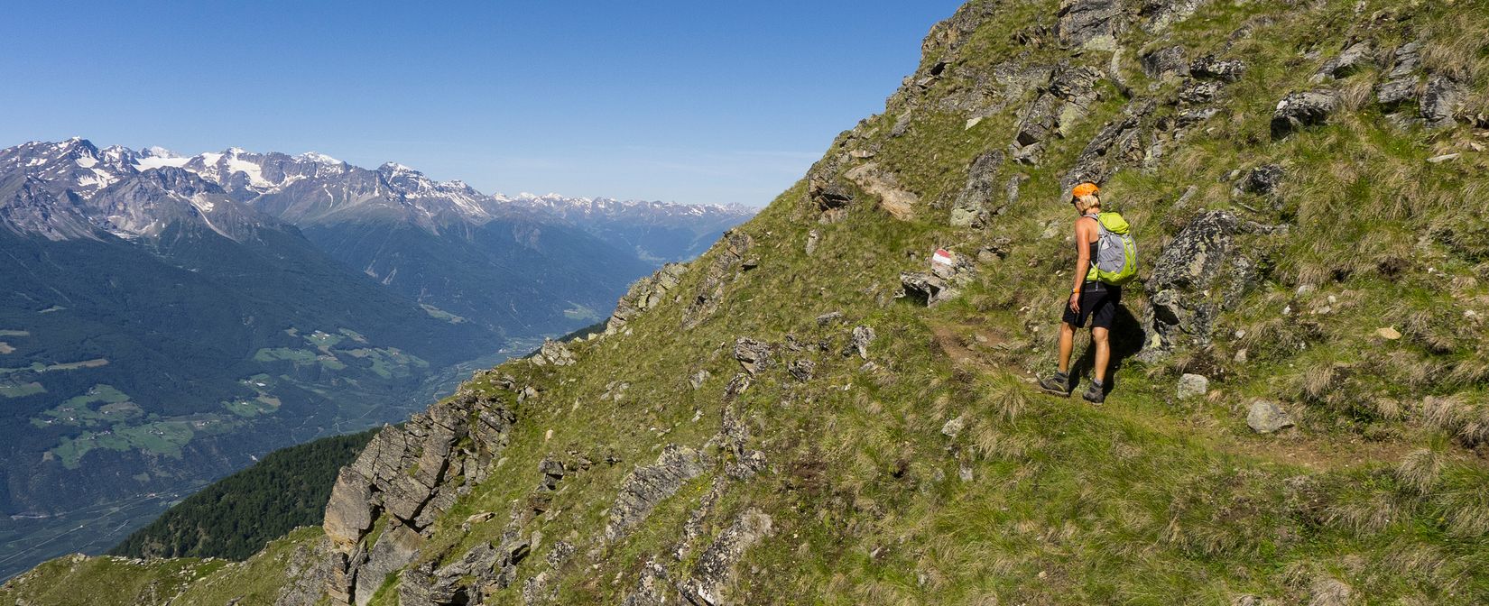 Eine Wanderin mit Rucksack und Helm geht auf einem schmalen Bergpfad an einem steilen, felsigen Hang entlang, während im Hintergrund ein weites Tal und schneebedeckte Gipfel zu sehen sind.